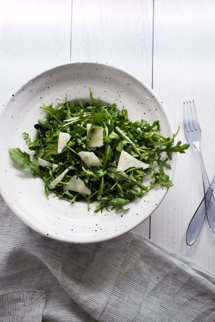 Arugula in a white bowl with shaved pecorino romano cheese on top next to a blue fork