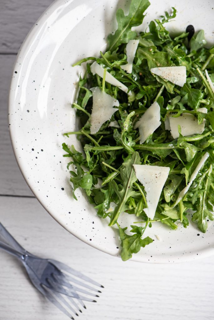 arugula salad greens in a white serving bowl with shaved Romano cheese on top